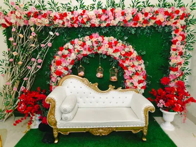 Elegant white and gold vintage sofa with vibrant red, pink, and white floral arch decoration and hanging candle lanterns for wedding or reception event in Behala, Kolkata