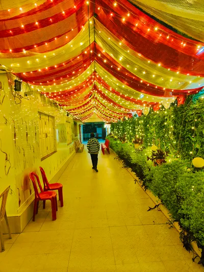 Decorated banquet hall corridor with red and yellow draped ceiling, string lights, green plants, and red chairs for wedding event in Behala, Kolkata
